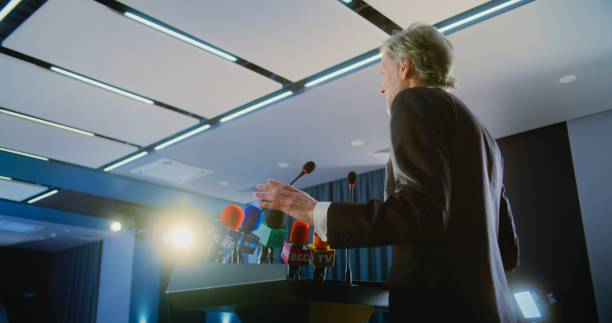 Female press officer pronounces speech from tribune during press campaign in the conference hall. Mature politician or activist answers questions, gives interview for television or media. Slow motion.