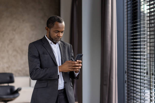 istock Concentrated black man in business suit holding cell phone in hands while standing next to window with jalousie. Attentive head of company solving business moments and making decisions at home 2148295191