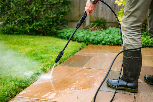 A senior man in his 70s cleans his patio stone slabs using a pressure washer. The man wears a green and blue check shirt with a green gilet.
