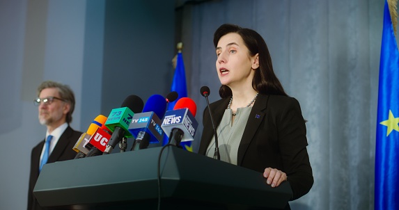 Female representative of the European Union during press conference. Confident politician makes an announcement, answers journalists questions and gives interview for media. Backdrop with EU flags.