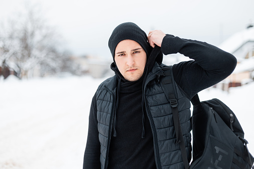 Handsome young stylish man in black fashionable dress with a vest, a hoodie, a hat and a backpack walks on the street on a winter snowy day