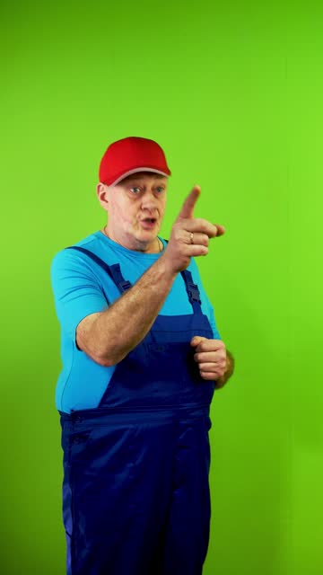 Senior repairman in overalls pointing someone with finger and inviting gesture.