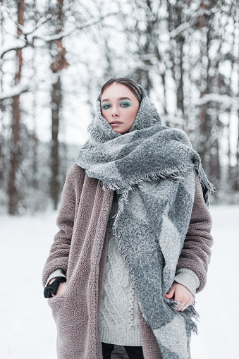 Russian beautiful young model woman in fashionable outerwear with vintage knitted scarf and fur retro coat posing in the woods with snow on a cold winter day