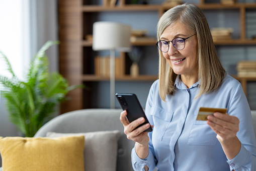 Mature woman holding credit card and smartphone, purchasing online from the comfort of home.