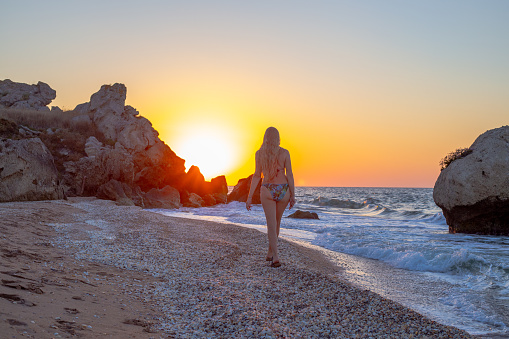 woman with long blond hair in a swimsuit walks along the rocky seashore at sunset. Outdoor recreation in summer.
