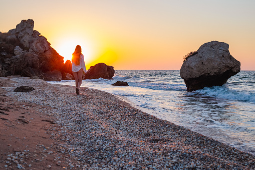 A woman with long blond hair walks along a rocky seashore at sunset. Outdoor recreation in summer.