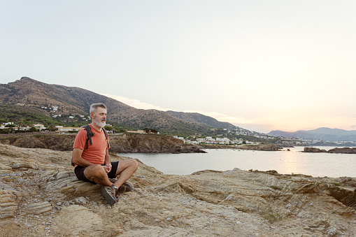 Adult man with a white beard sitting on some rocks meditating by the sea with his eyes closed.