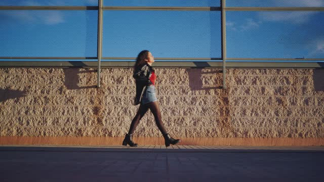 Young woman walking confidently along a wall with chain link fence at sunset