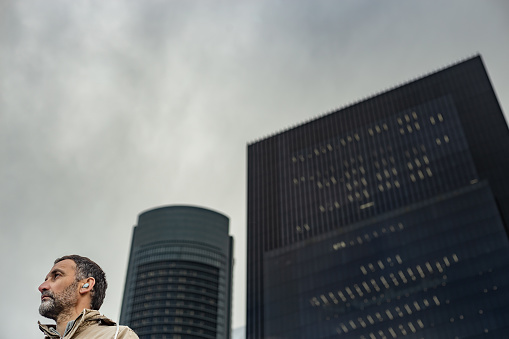 Madrid, Spain. The image depicts a scene where a mature man with in-ear headphones is seen from a low angle, with office skyscrapers in the background on a cloudy day. The man stands out for his defined profile and concentrated expression, indicating he is immersed in some activity. The in-ear headphones suggest he is listening to something privately. The office skyscrapers rise imposingly into the cloudy sky, conveying a sense of grandeur and power. The gray and cloudy atmosphere suggests a day filled with challenges or reflection.