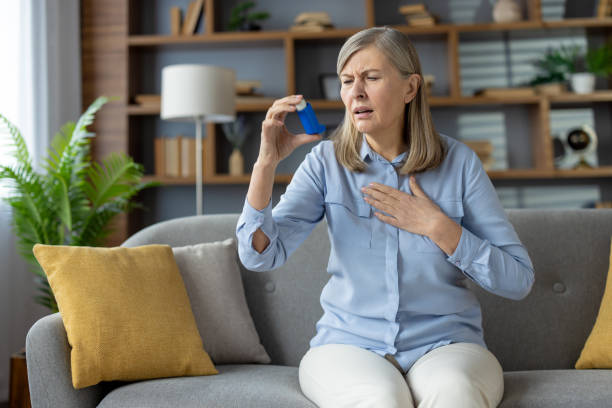 istock Grey haired woman in light outfit holding arm on chest while using medication for respiratory problem in living room. Disturbed patient experiencing asthma attack and relieving breathing struggles 2148301374