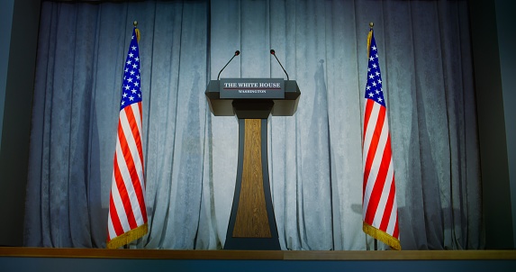 Tribune for politician, congressman or US minister political speech in the White House in press campaign room. Wooden conference debate stand with microphones on stage. American flags in background.