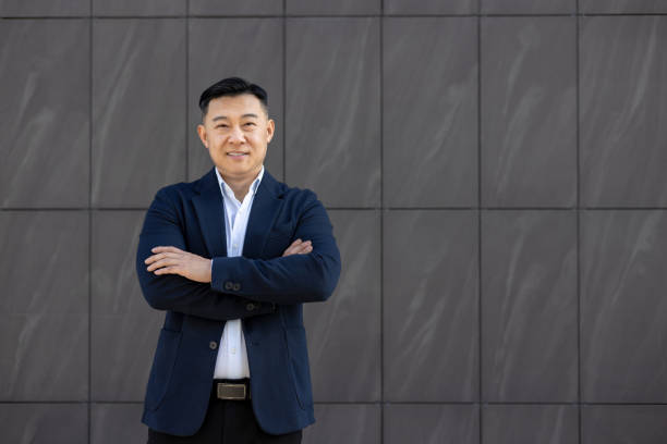 Portrait of young successful Asian businessman, lawyer in suit standing outside against wall background, arms crossed on chest and smiling confidently at camera Portrait of young successful Asian businessman, lawyer in suit standing outside against wall background, arms crossed on chest and smiling confidently at camera. asian suit man cross hand stock pictures, royalty-free photos & images