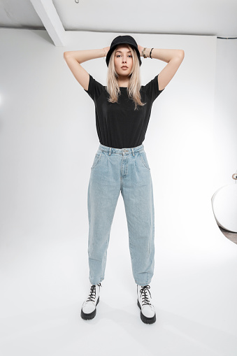 Stylish pretty young woman in fashionable clothes with blue jeans, black Panama hat, T-shirt and white shoes boots in the studio on a white background