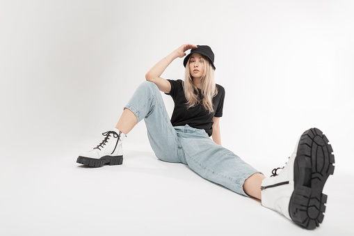 Beauty cool stylish young model girl in fashionable clothes with boots, blue jeans, a black T-shirt and Panama hat sits on a white background. Pretty woman with fashion shoes