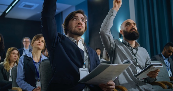 Journalists sit and write answers in notebooks from politician during press campaign in the conference hall. Caucasian male media representative raises his hand and asks questions during interview.