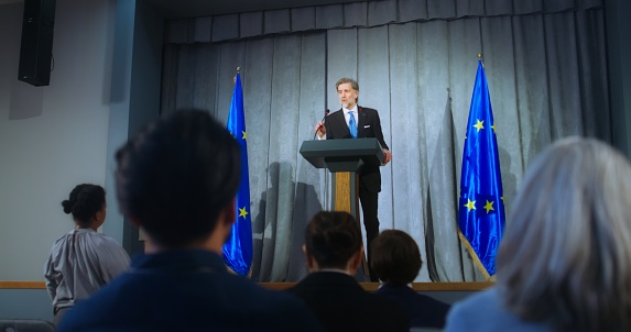 Mature representative of the European Union during performance at press conference. Confident politician makes an announcement, answers media questions and gives interview. Backdrop with EU flags.