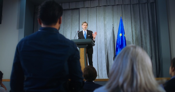 Mature representative of the European Union during performance at press conference. Confident politician makes an announcement, answers media questions and gives interview. Backdrop with EU flags.