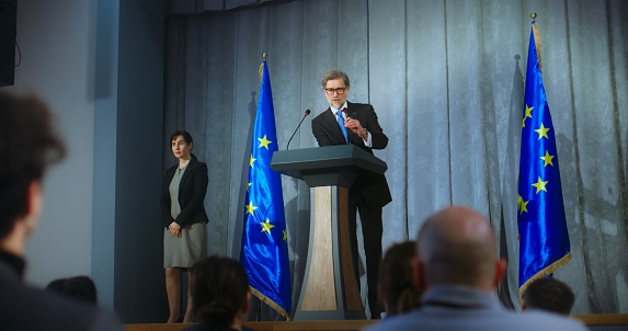 Mature politician comes to tribune, delivers campaign speech, answers journalists questions, gives interview to media. Representative of the European Union performs at press conference. Elections day.