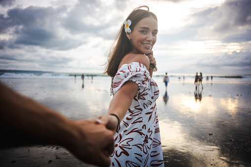 A young, beautiful white woman holding hands with her boyfriend during a sunset at Bali beach.
