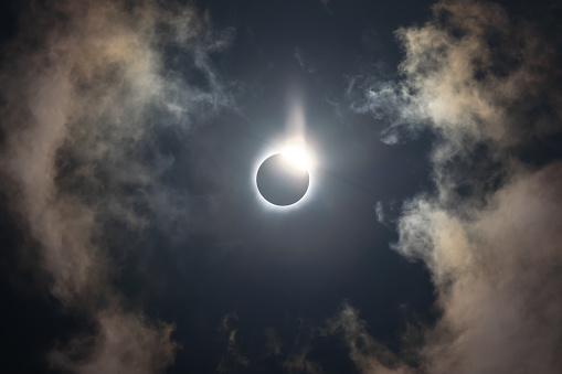 Total solar eclipse of April 8th 2024 with the sun peaking from behind the moon creating. a diamond ring effect. Totality with bright corona visible against dark blue sky with a few clouds.