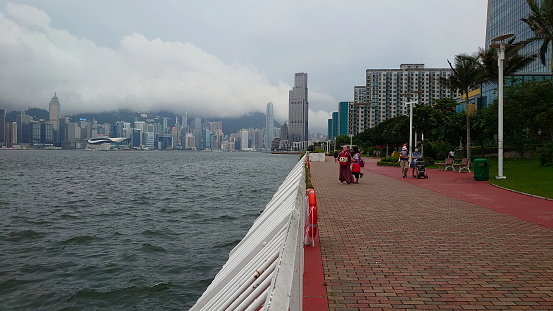 Hong Kong - 08.01.2021: Passengers walking on Hung Hom Promenade along the Victoria Harbour during Covid19 with Wai Chai in Hong Kong Island and Tsim Sha Tsui in Kowloon in the back under a cloudy sky
