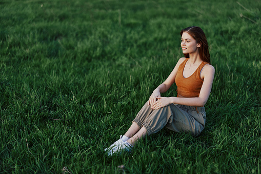 A happy woman looks out at the setting summer sun sitting on the fresh green grass in the park and smiling, view from above. The concept of self-care. High quality photo