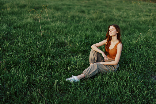 The redheaded woman sits in the park on the green grass wearing an orange top, green pants, and sneakers and looks out at the setting summer sun. High quality photo