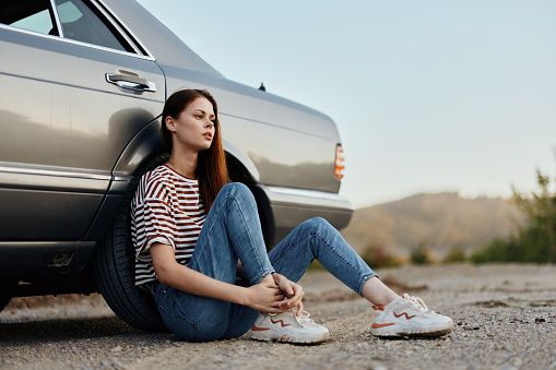 A young woman sits on the ground near her car on the side of the road and looks at the sunset. Camping after a hard road trip. High quality photo