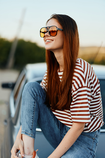 A fashionable woman smiles sweetly in stylish sunglasses, a striped t-shirt and jeans, sits on the trunk of a car and looks at the beautiful nature of autumn. Travel lifestyle. High quality photo