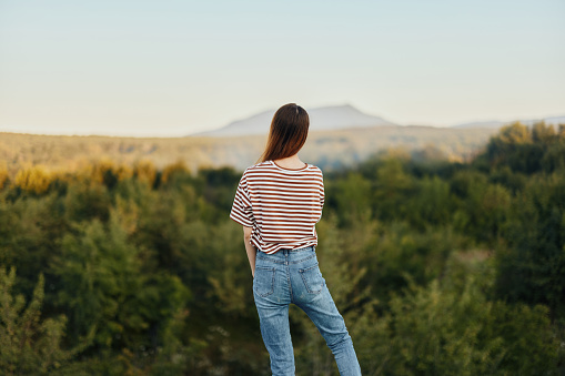 A young woman stands with her back to the camera in a T-shirt and jeans in nature and admires a beautiful view of the mountains. High quality photo