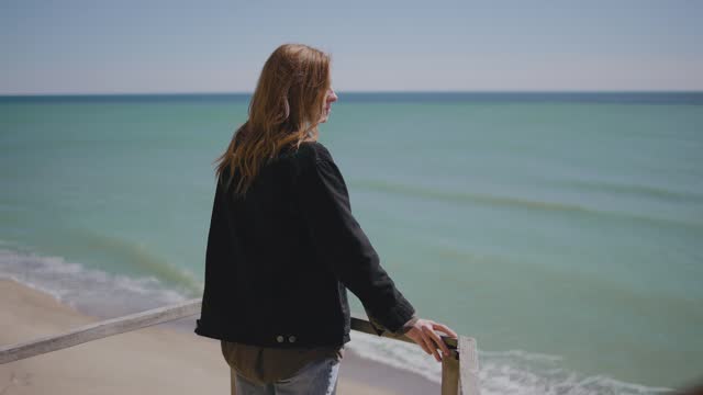 Attractive redhead woman in coat walking on the wild autumn beach. Woman goes down the wooden stairs to a deserted beach
