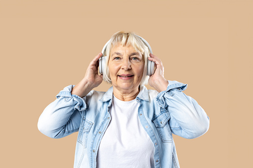 Cheerful elderly blonde woman lady 60s years old in casual clothes listening music with headphones isolated on pastel beige color background