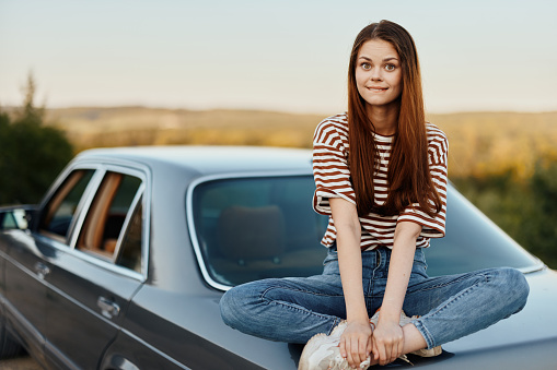 A young woman sits on the trunk of a car and rests after a difficult road and admires nature with a beautiful view. Stopping is also part of the journey. High quality photo