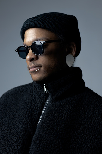 African-American male with black-framed tinted glasses, all black clothing, and left lip-piercing; looking away; jacket zippered up