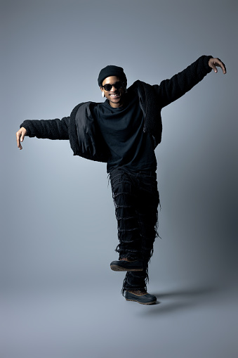 African-American male with black-framed tinted glasses, all black clothing, and left lip-piercing; smiling while standing on left leg with arms raised; jacket unzipped