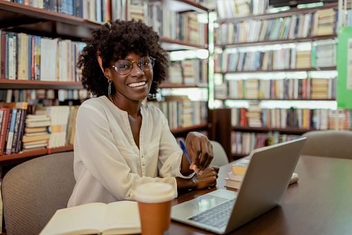 Shot of young satisfied black female student looking at camera while sitting at university library or book store