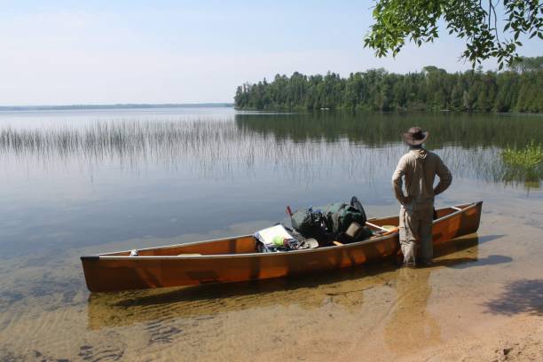 A couple paddles a red canoe on a serene lake, surrounded by lush, forested hills. The calm water reflects the clear blue sky, capturing the tranquility of nature and a sense of peaceful solitude.