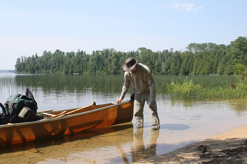 A man stands in shallow water as he unloads his canoe near the shore of a sandy beach in Quetico Provencial Park.