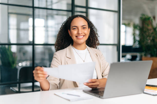 Pretty brazilian or hispanic business woman, company employee, financial ceo, hr manager, sits at a work desk in modern office, working in a laptop, studying financial documents, smiles at camera stock photo