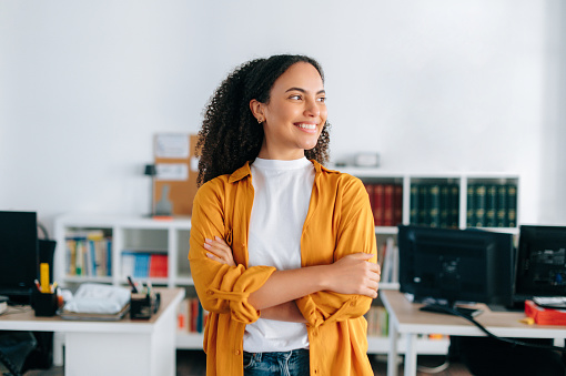 Amazing multiracial curly haired young woman in a casual orange shirt, freelancer, office employee, stand in the office near work desk with arms crossed, looks away, smiling happily
