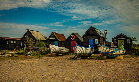 Boats moored outside a row of beach huts on a blue sky cloudy day.