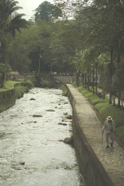 An elderly man with a cane in light clothes walks next to a canal in the daytime