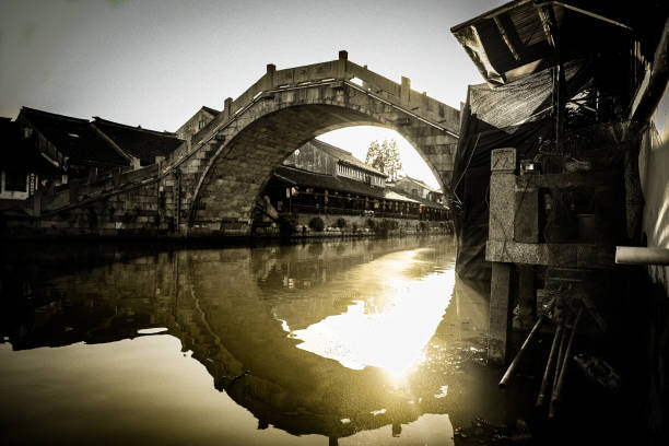A bridge in the center of the shot forms a ring with its reflection as sun reflects off the river water and traditional architecture lines both banks