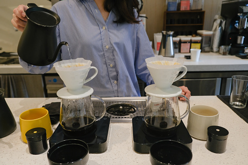 Close-up shot of unrecognizable Asian barista pouring hot water from kettle into coffee dripping filter above coffee maker jug