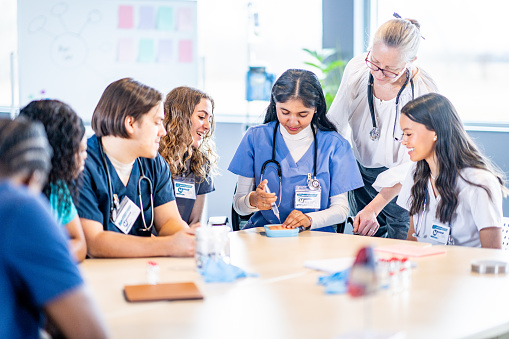A small group of medical students sit around a table in class as their instructor guides them through various procedures and closely supervises.  The students are each dressed professionally in scrubs and watching attentively.