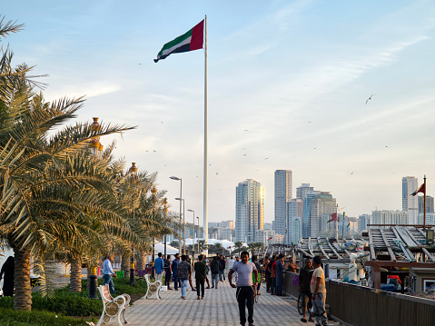 tourists and locals walk along the Sharjah waterfront against the background of the UAE flag. Sharjah, United Arab Emirates, January 28, 2024.