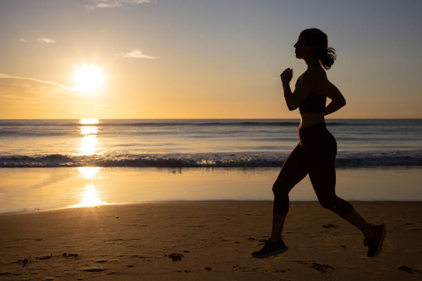 Woman at sunrise on the beach doing yoga or Pilates exercises, reaching up to the sky and being healthy and well at dawn and sunrise. Sun peaking over the clouds on a beautiful seafront. Female health and wellness on the beach, yoga, pilates, running, exercising and stretching in the morning. Happiness, fitness, movement, well-being at sunrise or sunset on the sand with crashing waves in the background. Golden hour sun rising over ocean. sunset dew stock pictures, royalty-free photos & images