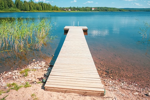 A wooden dock stretches out into a calm lake reflecting the blue sky and trees. The water is serene, mirroring the fluffy white clouds above. The scene exudes tranquility on a sunny day.