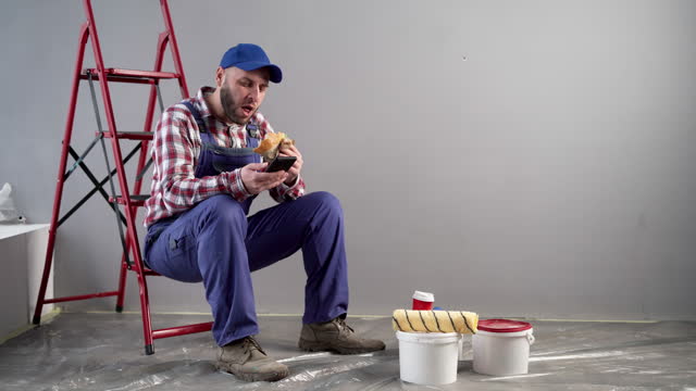Professional painter having lunch with burger uses mobile phone for chatting with friends, sitting on ladder. Decoration and improvement home interior