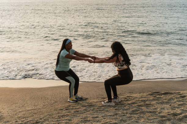 istock Latin women in workout attire hold hands while performing squats on a sandy beach 2148338629